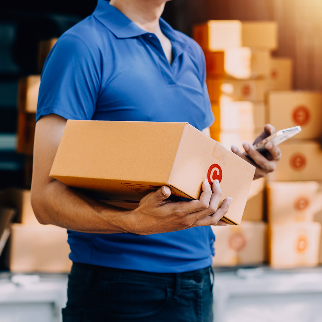 Person in a blue shirt holding a package and a smartphone in a warehouse.